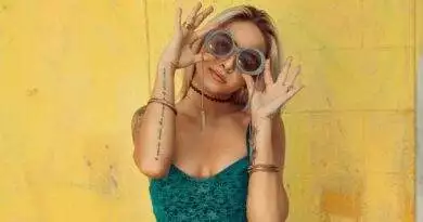 Eine Frau in einem trendigen grünen Top und Sonnenbrille posiert vor einer leuchtend gelben Wand.