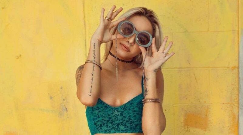 Eine Frau in einem trendigen grünen Top und Sonnenbrille posiert vor einer leuchtend gelben Wand.